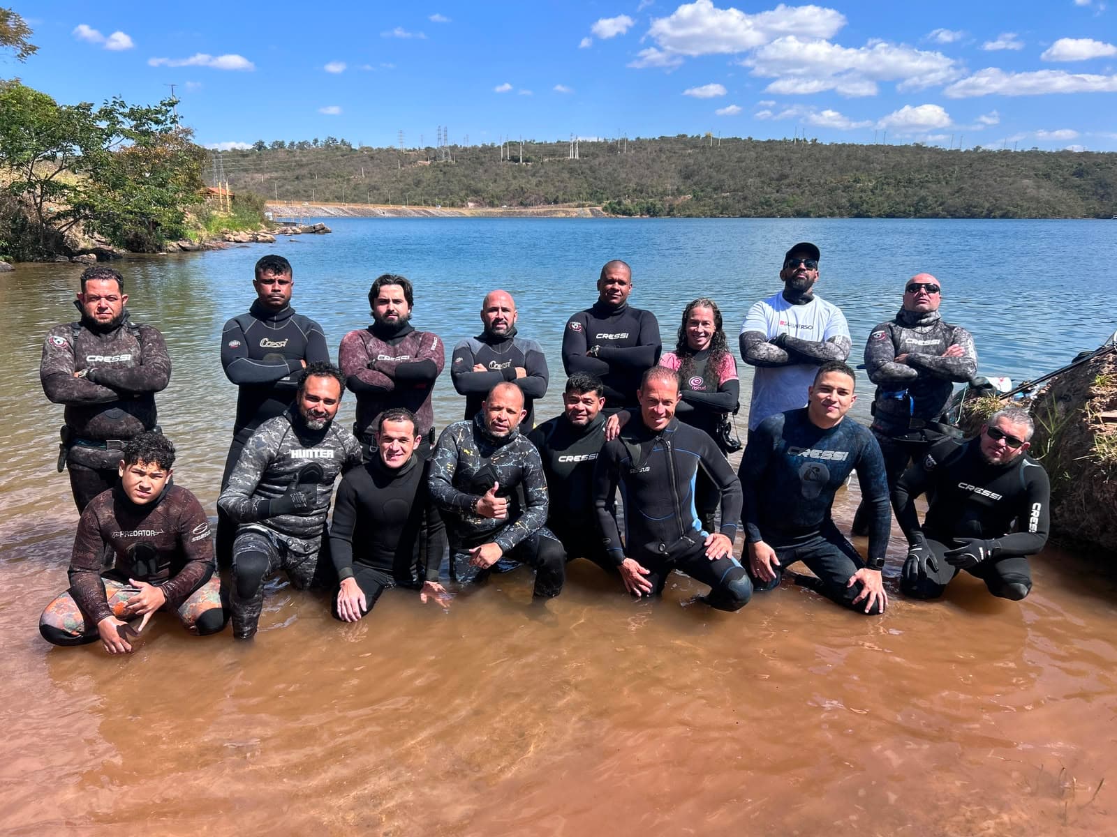 Turma de alunos em roupa de neoprene na beira da represa, ao fim de uma aula prática